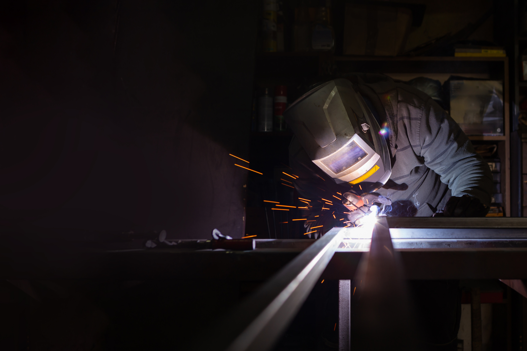 An adult male works in a workshop. He holds a welding machine in his hand and welds the metal structure.